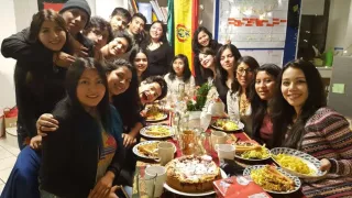 18 Freiwillgie aus Bolivien sitzen und stehen um einen Tisch. Sie blacken in die Kamera und lachen. Der Tisch ist mit Gläsern, Tassen, Kuchen, Spätzle und Würstchen gedeckt.