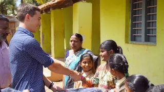 Ein Freiwilliger steht vor mehreren Kindern. Einem Mädchen reicht er die Hand. Im Hintergrund ist eine Frau und ein Mann zu sehen.