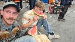 Zwei Männer sitzen auf dem Bordstein einer Straße. Der Linke von beiden lächelt in die Kamera und hält ein Stück Melone hoch, während der Rechte von beiden in eine Melone beißt und in die Kamera schaut.
