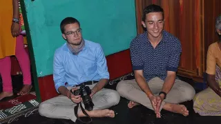 Zwei Freiwillige sitzen im Schneidersitz auf dem Boden. Der linke der beiden Männer hält eine Kamera in der Hand. Der Rechte faltet seine Hände zusammen und schaut auf eine Person gegenüber von ihm.