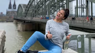 Lara is sitting on a wall on the bank of the Rhine with both thumbs up. In the background, a bridge across the Rhine is visible along with Cologne Cathedral on the opposite bank of the river.