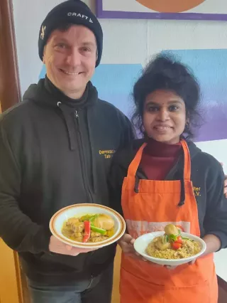 Sushmila steht neben einem Mann und beide halten einen Teller Essen in die Kamera.