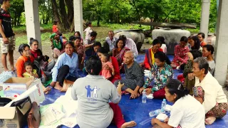 Eine Person der NGO sitzt mit einigen Menschen unter einem offenen Dach auf dem Boden. Die Person erklärt etwas. Die Menschen sitzen in Reihen und hören Ihr zu. Im Hintergrund sind grüne Bäume und Sträucher zu sehen.