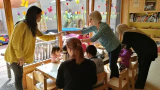 Les enfants sont assis avec leurs parents à une table sur laquelle est posé le repas. La table se trouve devant une baie vitrée, recouverte de papillons multicolores bricolés. Deux adultes se passent une tasse de café.