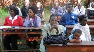 Männer und Frauen sowie ein kleines Kind aus Äthiopien sitzen an Schulbänken, die sich draußen im Freien auf einer Wiese befinden. Im Hintergrund an der letzten Schulbank sitzt der weltwärts-Freiwillige Paul, neben einer anderen Freiwilligen.