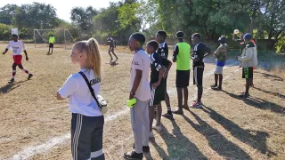 Acht Personen stehen am Seitenrand eines Fußballfeldes mit dem Rücken zur Kamera und beobachten ein Fußballspiel.