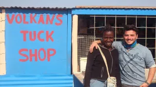 Ein junger Mann und eine junge Frau stehen nebeneinander vor einem kleinen Shop mit der Beschriftung „Volkans Tuck Shop“.