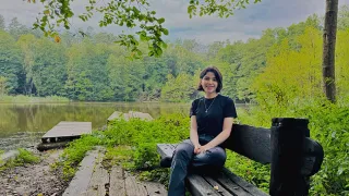 Eine Frau sitzt auf einer Bank im Wald und einem See im Hintergrund und lächelt in die Kamera.