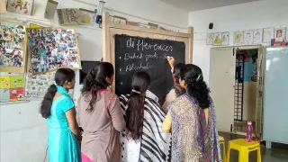 Fünf Frauen stehen vor einer einfacheren Schultafel in einem Klassenraum. Eine Frau schreibt mit Kreide in Englisch das Wort differences auf.