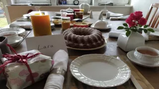 Una mesa decorada con velas, una tarta y rosas en un jarrón. En el sitio de Lara hay una tarjeta con su nombre y un regalo envuelto.