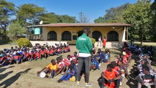 Viele Jungen sitzen in sieben Reihen hintereinander auf dem Boden. Vor ihnen steht ein Mann mit dem Rücken zur Kamera auf dessen Rücken die Aufschrift „Zambia“ zu lesen ist.