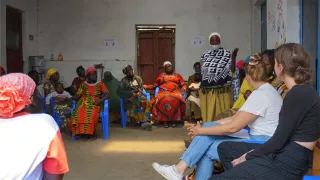 Some women from the project are sitting together in a circle. One woman is standing and appears to be explaining something. The other women are looking attentively at her. Leonie and her fellow volunteers are sitting and listening to the woman.