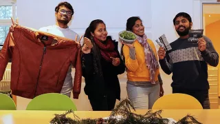 Four volunteers are standing next to each other, each holding an object. The objects are a leather jacket, a loaf of bread, rocket leaves and coffee.