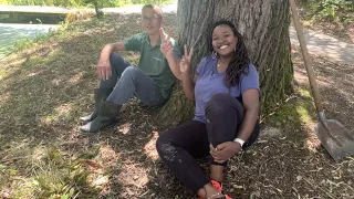 Elizabeth sitzt mit ihrem Kollegen angelehnt an einen großen Baum auf dem Boden und lächelt in die Kamera. Rechts im Bild ist eine Schaufel zu sehen.