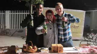 Drei junge Männer hinter einem Marktstand. Sie haben ein Bier in der Hand und halten dies vor sich in Richtung Kamera.