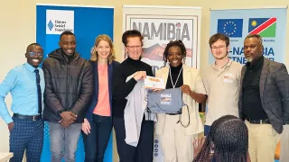 Sieben Personen stehen nebeneinander und lächeln in die Kamera. Zwei Frauen in die Mitte halten ein Schild mit der Aufschrift „Namibia needs me“ hoch.