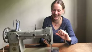 Eine Frau lacht in die Kamera und hat vor sich eine Nähmaschine auf einem Tisch stehen. Sie versucht einen Faden mit ihren Händen in die Maschine einzufädeln.