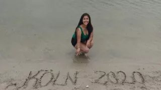 Lara is kneeling on the shore of a lake, pointing at the words “Köln 2020” (“Cologne 2020”) written in the sand.
