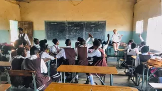 Circa 15 Kinder sitzen mit dem Rücken zur Kamera in einem Klassenraum. Einige zeigen auf, während die Lehrperson spricht.