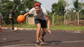 Der Freiwillige steht gebeugt mit einem Basketball in der rechten Hand. Im Hintergrund spielt ein Kind mit einem Fußball.