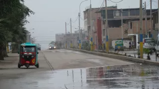 On aperçoit une rue. On voit une voiture au loin. À gauche sur le bord se trouve un moto-taxi. À droite, on distingue des poteaux électriques et des maisons.
