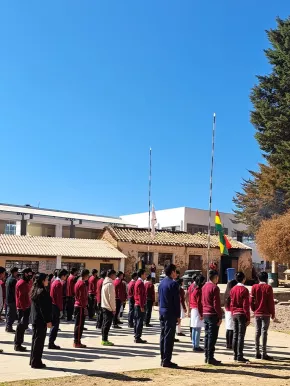 Über 20 Kinder stehen in roten Schuluniformen mit dem Rücken zur Kamera. Sie schauen alle nach vorne, wo die rot-gelb-grüne Flagge von Bolivien an einem Pfahl gehisst wird.
