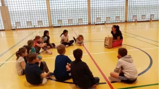 Children are sitting in a circle with Lara in a gym. The children are listening to Lara.