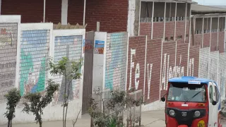 La photo montre un bâtiment en briques avec de hauts murs. Les murs sont peints et portent le nom de l'école, « Fe y Alegría ». Devant l'école, on distingue de petits arbres et un moto-taxi.