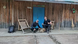 Zu sehen sind zwei Männer, welche vor einer Holzhütte sitzen. In der Mitte der beiden ist ein Hund zu sehen, welcher von einem der beiden gestreichelt wird.
