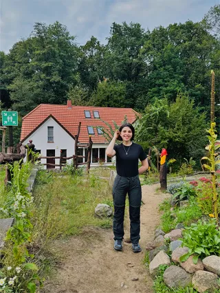 Eine Frau steht lächelnd auf einem Weg, im Hintergrund ein ländliches Haus und Wald.