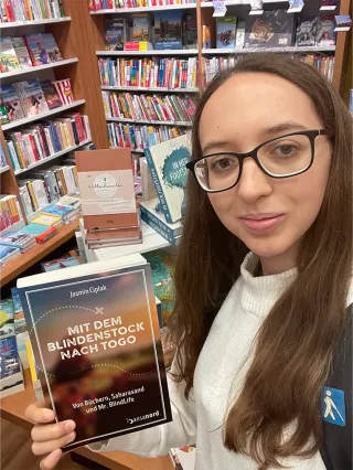 Jasmin Ciplak steht in einer Buchhandlung, schaut in die Kamera und hält in der einen Hand ihr Buch.