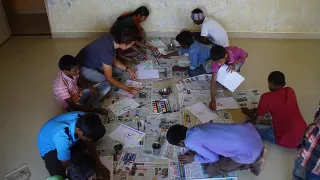 Mehrere Kinder knien auf dem Boden und malen mit Wasserfarbe und Pinsel auf weißes Blattpapier. Ein Freiwilliger und eine indische Mitarbeiterin knien ebenfalls auf dem Boden und sprechen mit Kindern.