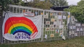 A large cloth with a self-painted rainbow and the words "Bleibt gesund" hangs on a grille. To the right of it hang small drawings of rainbows.