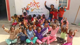 Eine Gruppe Kinder sitzt vor einer Steinmauer mit dem Spruch „DRC Group Project“ auf dem Boden. Die Kinder haben die Arme in die Luft gestreckt.