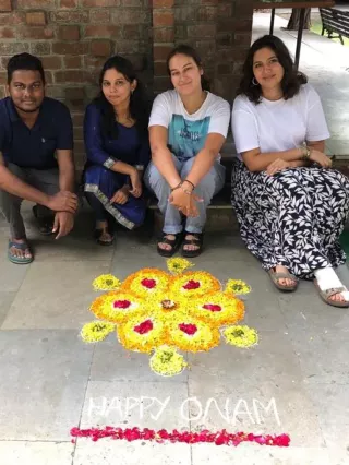 Vier Menschen sitzen auf dem Boden. Vor ihnen liegt ein Muster aus Blumen und die Aufschrift "Happy Onam"