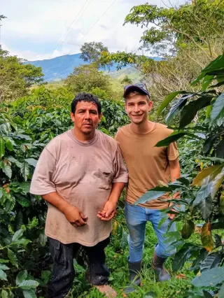 Zwei Männer stehen auf einer Kaffeefarm und lächeln in die Kamera.