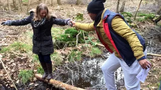 Une volontaire se tient sur un tronc d'arbre dans une forêt au-dessus d'un ruisseau. Un deuxième volontaire lui tend la main pour traverser le ruisseau.