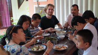 Der Freiwillige Simon sitzt bei seiner Einsatzstelle mit Kolleginnen und Kollegen am Essenstisch und isst Reis und Gemüse mit Stäbchen.