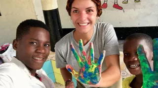 Eine Freiwillige steht neben zwei Kindern. Alle drei strecken ihre mit Wasserfarbe bemalte Hand in die Kamera.