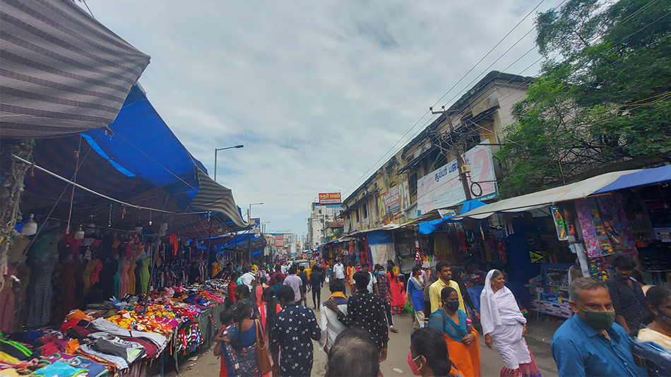 Links und rechts im Bild sind Marktstände zu sehen, an denen bunte Kleidungsstücke zu kaufen sind. In der Mitte verläuft die Einkaufsstraße, auf der einige Menschen unterwegs sind.