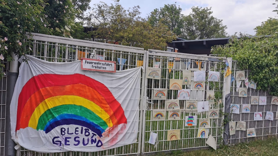 The fence of the school grounds is decorated with pictures and a large sheet painted with a colorful rainbow.