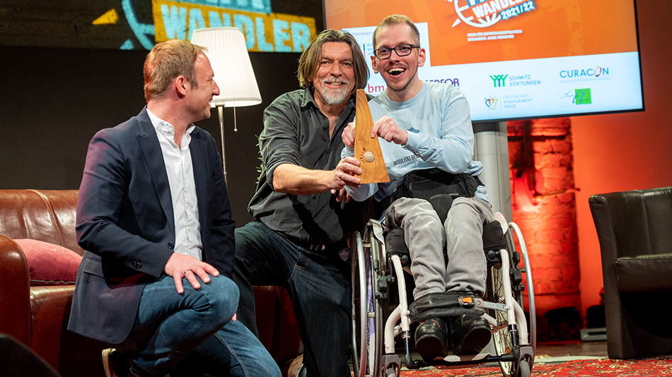 Drei Männer schauen in die Kamera. Preisträger Kevin Kleiber sitzt im Rollstuhl rechts im Bild und zeigt fröhlich den Preis in die Kamera.