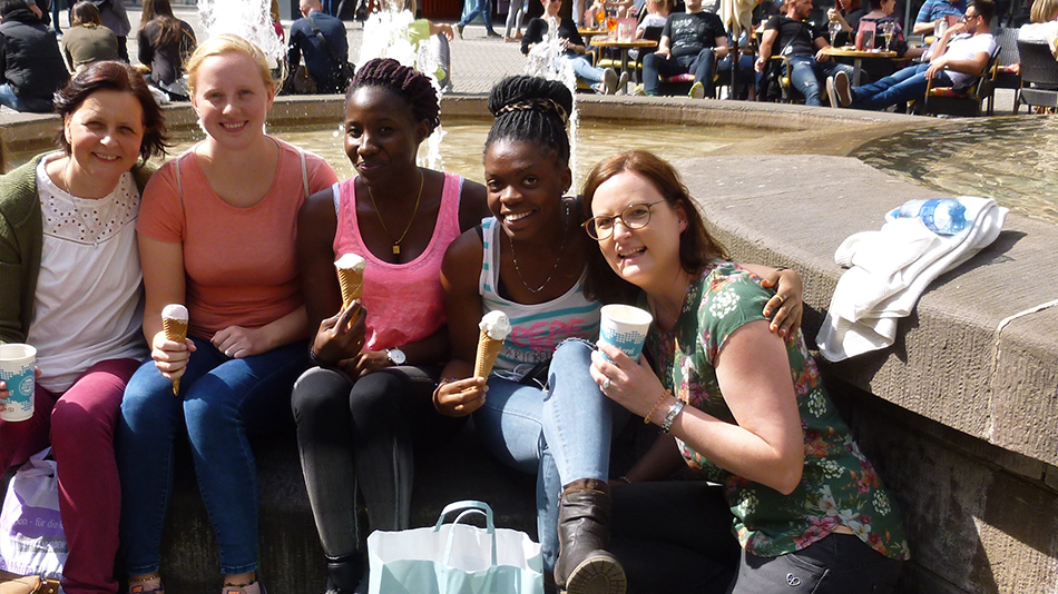 Beate, Carolin, Arijenida, Sandra and another woman sit in front of a fountain, eat ice cream and smile into the camera.