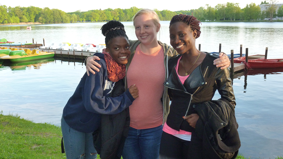 Sandra, Arijenida and Carolin smile into the camera. Carolin is in the middle and puts both arms around the other two women.