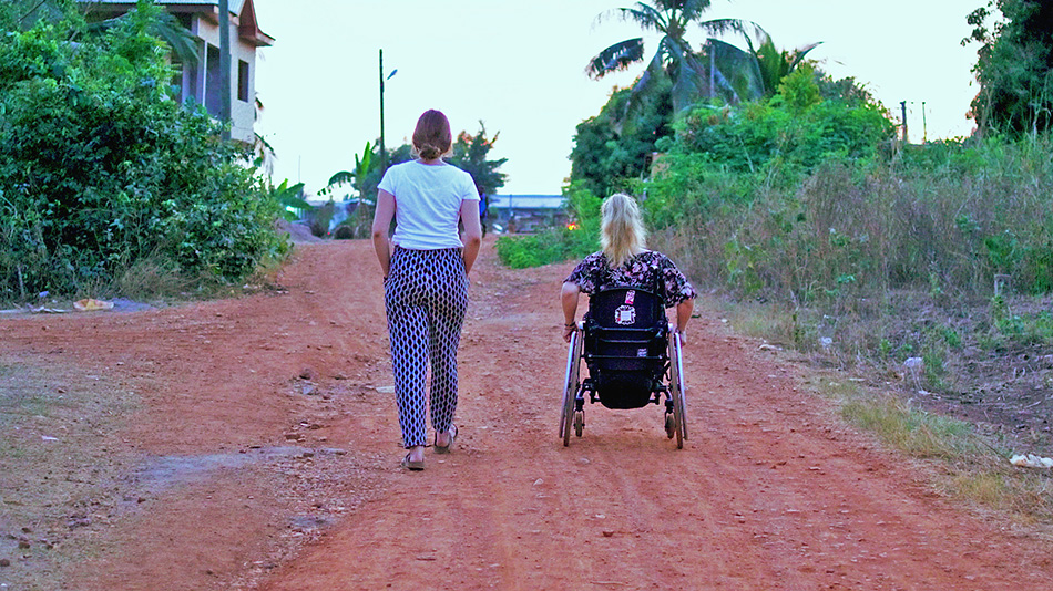 Auf dem Bild ist die weltwärts-Freiwillige Steffi zu sehen, die im Rollstuhl sitzt und gemeinsam mit einer anderen Person einen ungepflasterten Weg fährt,.