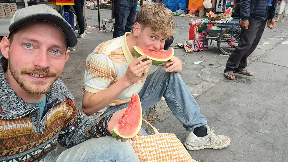 Zwei Männer sitzen auf dem Bordstein einer Straße. Der Linke von beiden lächelt in die Kamera und hält ein Stück Melone hoch, während der Rechte von beiden in eine Melone beißt und in die Kamera schaut.