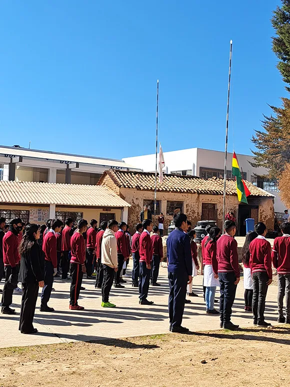 Über 20 Kinder stehen in roten Schuluniformen mit dem Rücken zur Kamera. Sie schauen alle nach vorne, wo die rot-gelb-grüne Flagge von Bolivien an einem Pfahl gehisst wird.