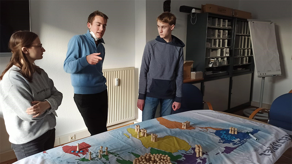 Trois jeunes gens se tiennent devant une table sur laquelle est installé un « jeu du monde ».